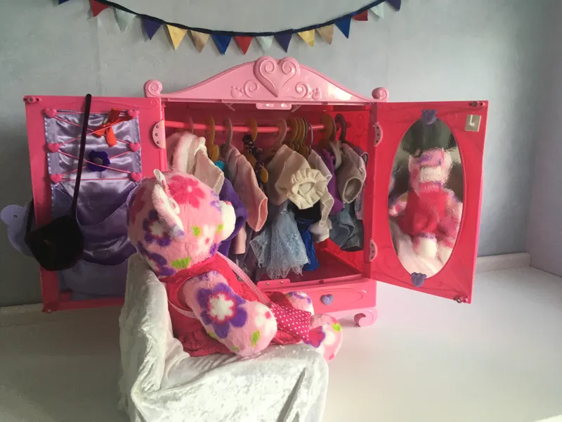 A pink floral bear seen from behind, looking at her reflection in the wardrobe mirror while holding up an outfit