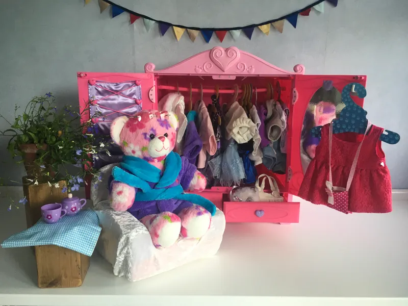 A pink floral bear in a turquoise dressing gown sitting on a little bed next to an open wardrobe
